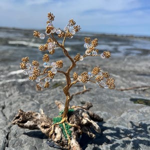 May include: A decorative wire tree sculpture featuring a twisted gold-coloured metal trunk and branches adorned with small, clear, iridescent beads. The base of the tree is anchored into a piece of textured, weathered driftwood with green moss.