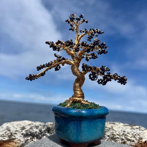 Peut inclure: Une sculpture artisanale de bonsaï avec un tronc et des branches en fil d'or, ornées de perles bleu foncé. L'arbre est dans un petit pot carré en céramique turquoise avec une base de mousse, sur fond de ciel bleu et d'océan.