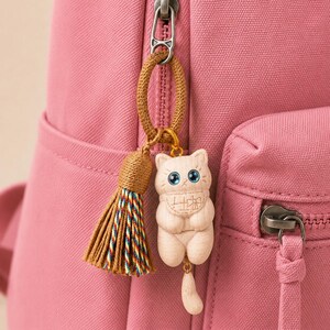 May include: A pink fabric backpack featuring a silver metal zipper pull attached to a brown cord loop. A decorative charm hangs from the loop, consisting of a brown tassel with multicolored threads and a light-colored wooden cat figurine.