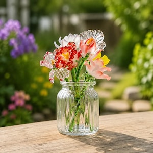 May include: A clear glass vase with vertical ridges contains a collection of colourful glass flowers. The flowers feature red, white, pink, and yellow petals. The vase is on a wooden surface, with a blurred garden backdrop.