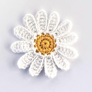 May include: A close-up of a crocheted daisy flower. The petals are white, and the centre is yellow. This handmade item could be used to decorate clothing, bags, or other craft projects. The daisy is a popular motif in crafts and represents purity.