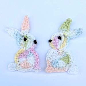 May include: Two crocheted pastel bunny rabbits with black eyes. The bunnies are facing each other and are made with a variety of pastel colours, including pink, blue, yellow, and green.