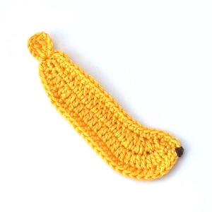 May include: A handmade, yellow crochet banana with a brown tip and stem. The banana is crafted with a textured stitch, showcasing the details of the yarn. The banana is set against a white background.