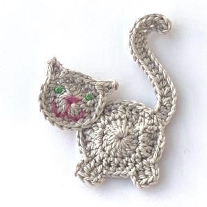 May include: A crocheted cat applique in shades of gray, featuring green eyes and a pink mouth. The cat has a curved tail and is made with a textured yarn. This handmade item is perfect for adding to clothing or accessories.