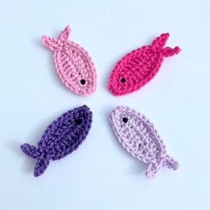 May include: Four crocheted fish in shades of pink, purple, and lavender, arranged in a circular pattern. Each fish has a small black eye and detailed fins. The handmade fish are set against a white background.