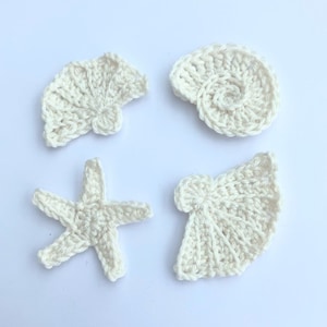 May include: Four cream-coloured crochet sea-themed shapes: a starfish, a spiral shell, and two fan-shaped shells. The handmade items are arranged on a white background. The crochet stitches are visible, giving a textured appearance.