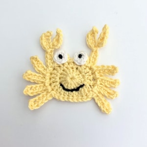 May include: A handmade crocheted crab applique in pale yellow. The crab features white and black eyes and a smiling mouth. The claws and legs are also yellow. Ideal for adding to clothing or other craft projects.