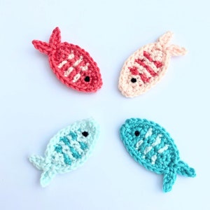 May include: Four crocheted fish in shades of red, peach, light blue, and teal. Each fish has a white pattern and a small black eye. The fish are arranged in a circular pattern on a white background.