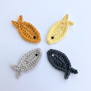 May include: Four crocheted fish arranged in a circle. The fish are in shades of yellow, gold, grey, and dark grey. Each fish has a small black or white eye. The fish have a simple, handmade appearance.
