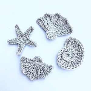 May include: Four crocheted sea-themed decorations in a silver colour. The decorations include a starfish, a seashell, and two different types of clams.