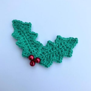 Könnte beinhalten: Ein grünes, gehäkeltes Stechpalmenblatt mit drei roten Perlen in der Mitte.