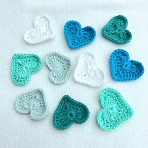 May include: A selection of crocheted heart shapes in shades of blue, green, and white. The hearts are displayed on a white surface. Each heart has a detailed, textured design, highlighting the handmade quality of the craft.