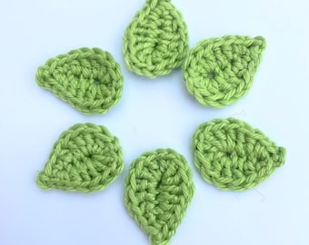 May include: Six crocheted green leaves, each with a small hole in the center, arranged in a circle on a white background.