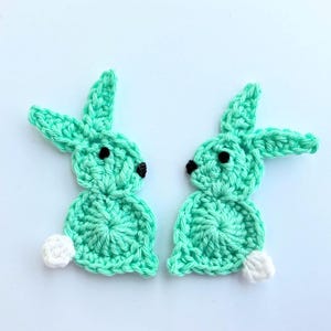 May include: Two crocheted green bunny rabbit shapes with white tails. The bunnies have black eyes and are facing each other.