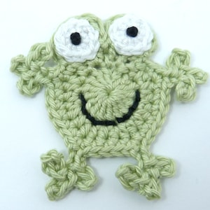 May include: A crocheted frog applique in light green yarn. The frog has large white eyes with black pupils and a smiling black mouth. The applique is handmade and has a textured appearance.