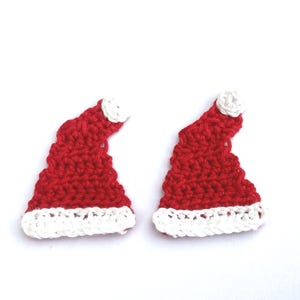 May include: Two handmade, red crochet Santa hats with white trim and a white pom-pom. The hats are set against a white backdrop. Ideal for festive crafting or embellishments, these hats are a charming seasonal item.