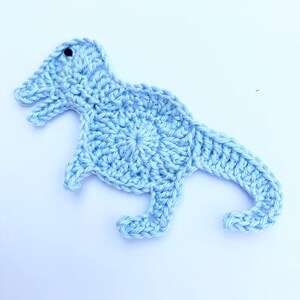 Puede incluir: Una aplicación de dinosaurio de crochet azul claro. El dinosaurio está de pie sobre un fondo blanco.