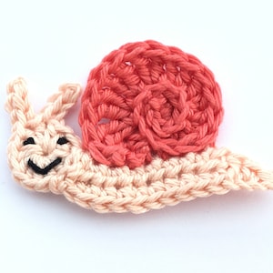 Peut inclure: Une applique en forme d'escargot crocheté, de couleur pêche et corail. Le corps de l'escargot est de couleur pêche clair, avec une coquille en spirale corail. L'escargot a un visage souriant avec des yeux noirs et une bouche noire.