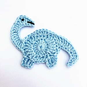 Puede incluir: Una aplicación de dinosaurio de crochet azul claro. El dinosaurio tiene un cuello largo y una cabeza pequeña con ojos negros.