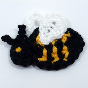 May include: A crocheted bee motif, featuring a black body with yellow stripes and white wings. The bee has small black antennae. This handmade item is likely intended for use in crafts or as an embellishment.
