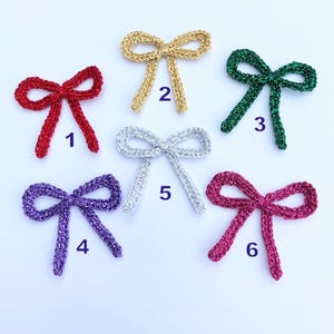 May include: Six crocheted bows in different colours, including gold, silver, red, green, purple, and pink. Each bow is numbered 1 through 6.