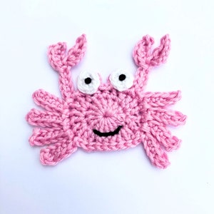 May include: A pink crochet crab with white and black eyes and a smiling mouth, set against a white background. The crab features detailed claws and legs. This handmade item is ideal for craft projects and adds a playful touch.