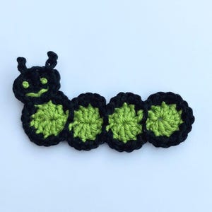 May include: A handmade crochet caterpillar in black and green. The caterpillar has a smiling face and is made of four connected circles. The body is green, and the edges are black. The antennae are black.