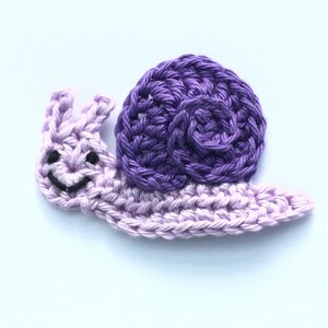 Peut inclure: Un escargot crocheté en violet et violet clair avec un visage souriant.