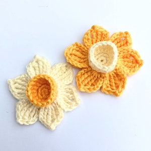 May include: Two handmade crochet daffodil flowers. One is cream and orange, the other is yellow and orange. The flowers have detailed petals and a central cup-shaped corona.