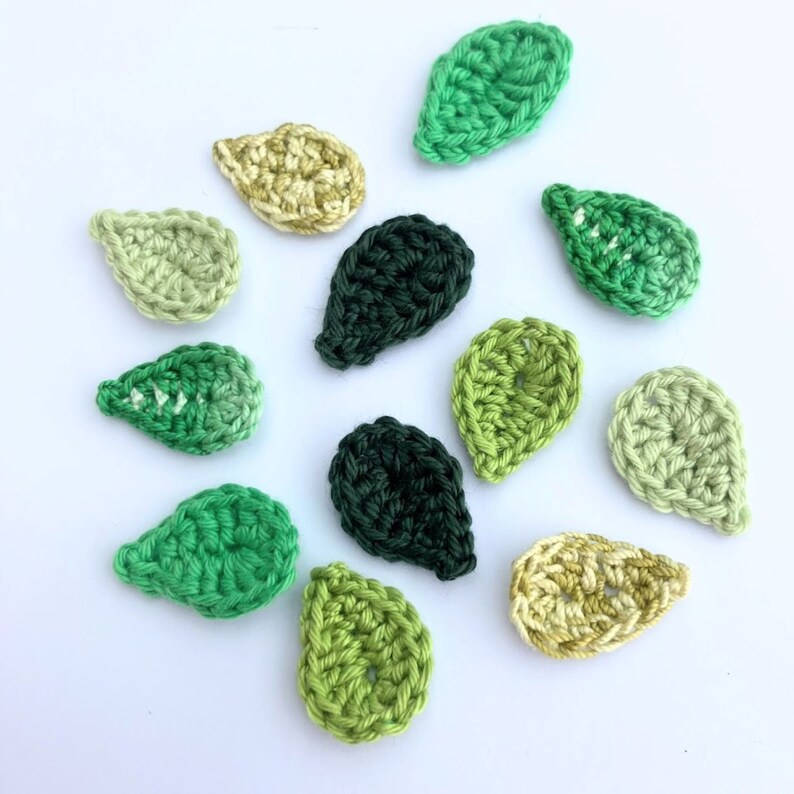 May include: A collection of crocheted leaf appliques in various shades of green and yellow. The leaves are handmade with a textured stitch, and arranged on a white surface. These decorative leaves are ideal for crafting projects.