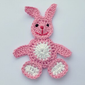 May include: A pink crocheted bunny with white accents. The bunny has a white circle in the centre of its body and white circles on its feet.