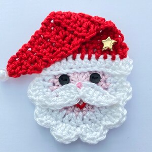 May include: A crocheted Santa Claus face with a red hat, white beard, and rosy cheeks. The hat has a gold star embellishment and a white pearl at the tip. Black button eyes and a red stitched mouth complete the design. A festive holiday decoration.