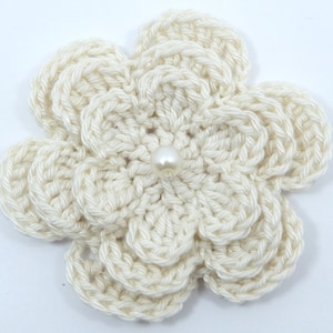 May include: A crocheted flower with a pearl in the centre. The flower is off-white and has five petals.