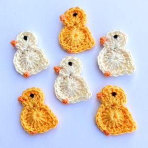 May include: Six handmade crochet ducklings in shades of yellow and cream. Each duckling has an orange beak and feet, and a black eye. The ducklings are arranged on a white surface.
