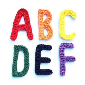 May include: A set of six crochet letters arranged in two rows. The top row features a red letter A, an orange letter B, and a yellow letter C. The bottom row displays a green letter D, a dark blue letter E, and a purple letter F, all crafted with yarn.