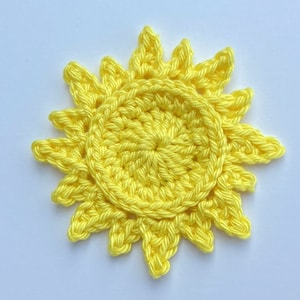 May include: A handmade, bright yellow crochet sun. The sun has a circular centre with radiating points, resembling sun rays. The craftwork is detailed, with visible stitches and a textured appearance. The item is likely a decorative element or applique.