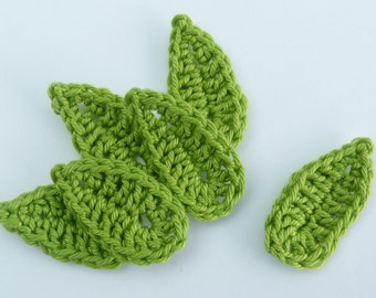 May include: A collection of six handmade, crochet leaves in a vibrant, light green color. The leaves are arranged on a white surface, showcasing their detailed stitch work and organic shape. These decorative elements are ideal for crafting projects.