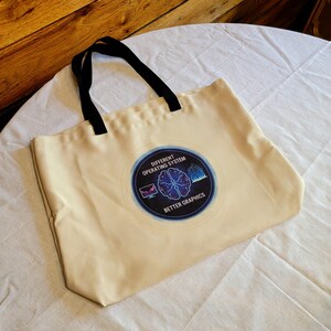 May include: Cream-colored tote bag with black handles. The bag features a circular graphic with the text "Different Operating System Better Graphics" and a brain design. The bag is made of a sturdy fabric.