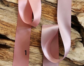 May include: Two samples of pink satin ribbon, one is a darker shade of pink and the other is a lighter shade of pink. The ribbon is displayed on a wooden surface. The ribbon is labeled with the numbers 1 and 2.