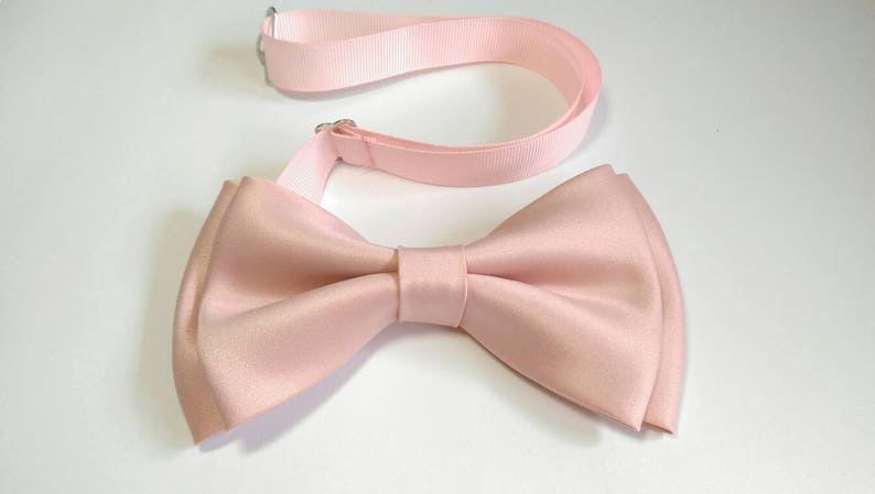 May include: A light pink bow tie with an adjustable strap. The bow tie is made of satin and has a classic shape.