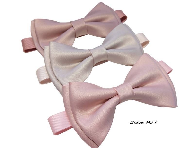 May include: Three satin bow ties in shades of pink and beige. The bow ties are arranged in a stack, with the lightest shade on top and the darkest shade on the bottom. The bow ties are tied with a ribbon and have a "Zoom Me!" label.