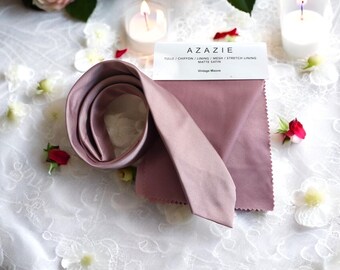 May include: A swatch of mauve fabric labeled "Vintage Mauve" with the text "Tulle/Chiffon/Lining/Mesh/Stretch Lining - Matte Satin" and the brand name "Azazie". A rolled up piece of the same fabric is also shown.