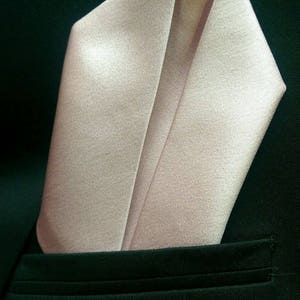 May include: A light pink satin pocket square folded in a classic point shape, against a dark green suit jacket.
