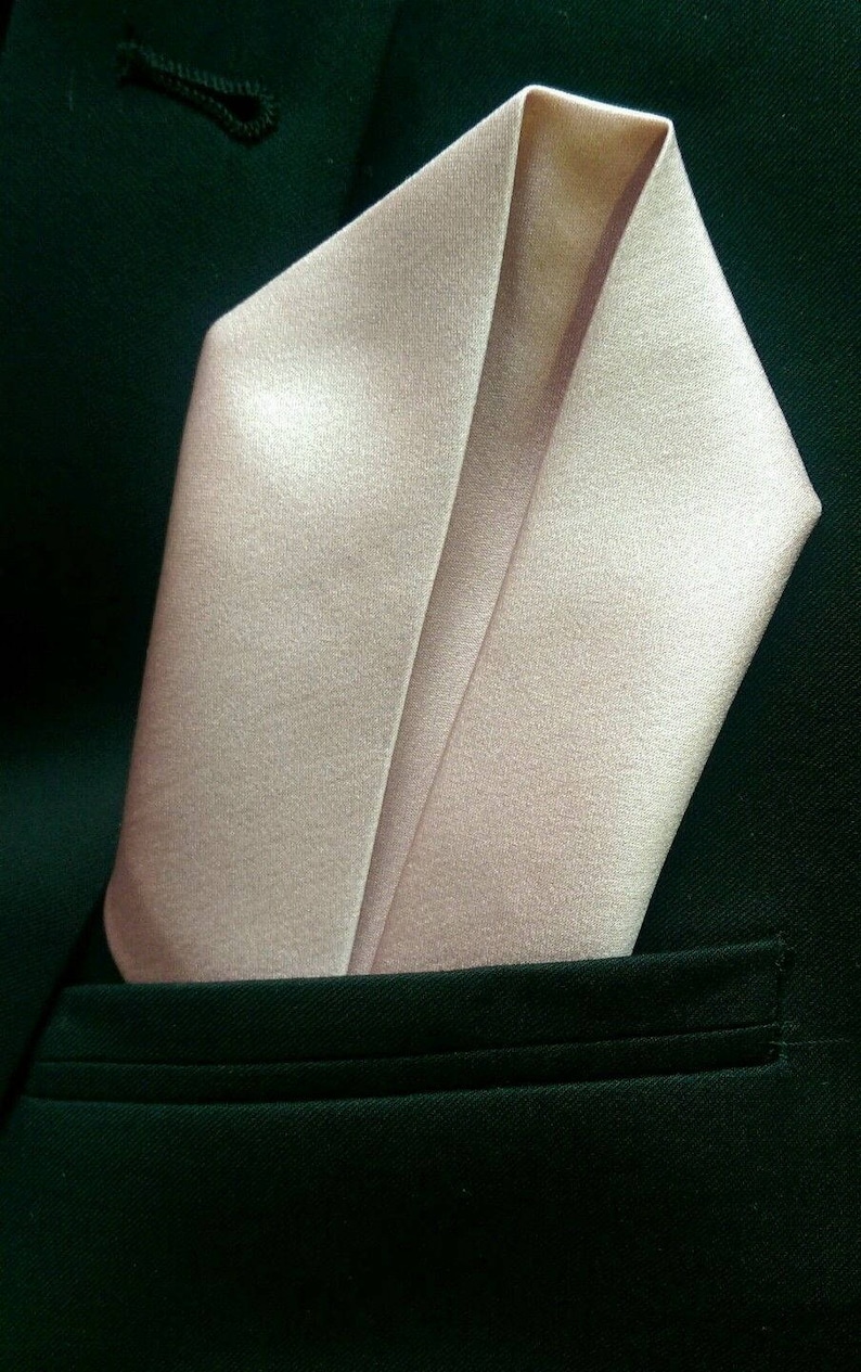 May include: A light pink satin pocket square folded in a classic point shape, against a dark green suit jacket.