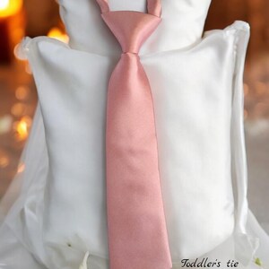 May include: A light pink satin toddler's tie. The tie is on a white pillow with white flowers. The text "Toddler's tie Light Rose" is below the tie.