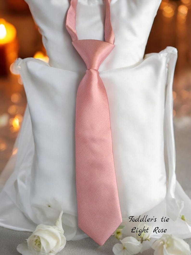 May include: A light pink satin toddler's tie. The tie is on a white pillow with white flowers. The text "Toddler's tie Light Rose" is below the tie.