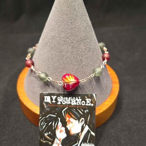May include: A beaded bracelet featuring a central red heart-shaped bead, alternating with smaller frosted green and red beads on a silver-toned chain. A square charm displays the My Chemical Romance Three Cheers for Sweet Revenge album cover.