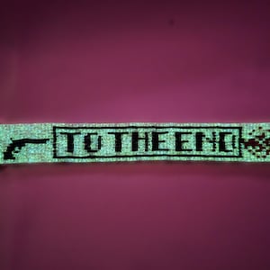 May include: A rectangular beaded bracelet featuring a light-reflecting surface with a black graphic of a handgun on the left, the text TO THE END in a black rectangular border, and a scattered pixelated effect on the right side with a metal clasp.