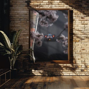 Puede incluir: Una obra de arte enmarcada que representa un coche de carreras en una pista, con flores de cerezo de fondo. La obra está colgada en una pared de ladrillo, con una ventana y una planta en maceta en primer plano.