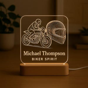 Może przedstawiać: Podświetlany znak akrylowy z motocyklem i kaskiem. Znak zawiera imię "Michael Thompson" i frazę "BIKER SPIRIT". Podstawa wykonana jest z drewna.